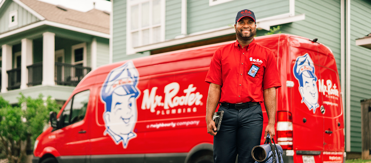 Mr. Rooter service professional carrying mat and utility bag in front of branded red work van.