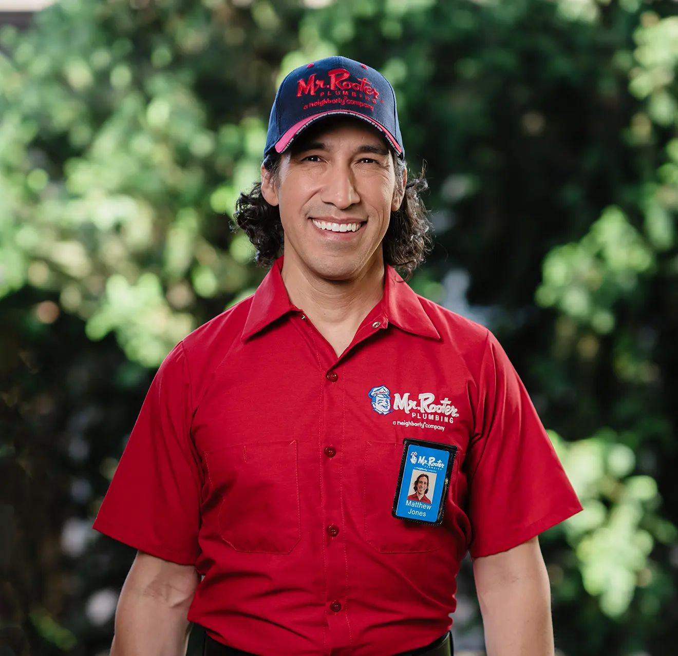 Smiling Mr. Rooter technician with greenery in the background.