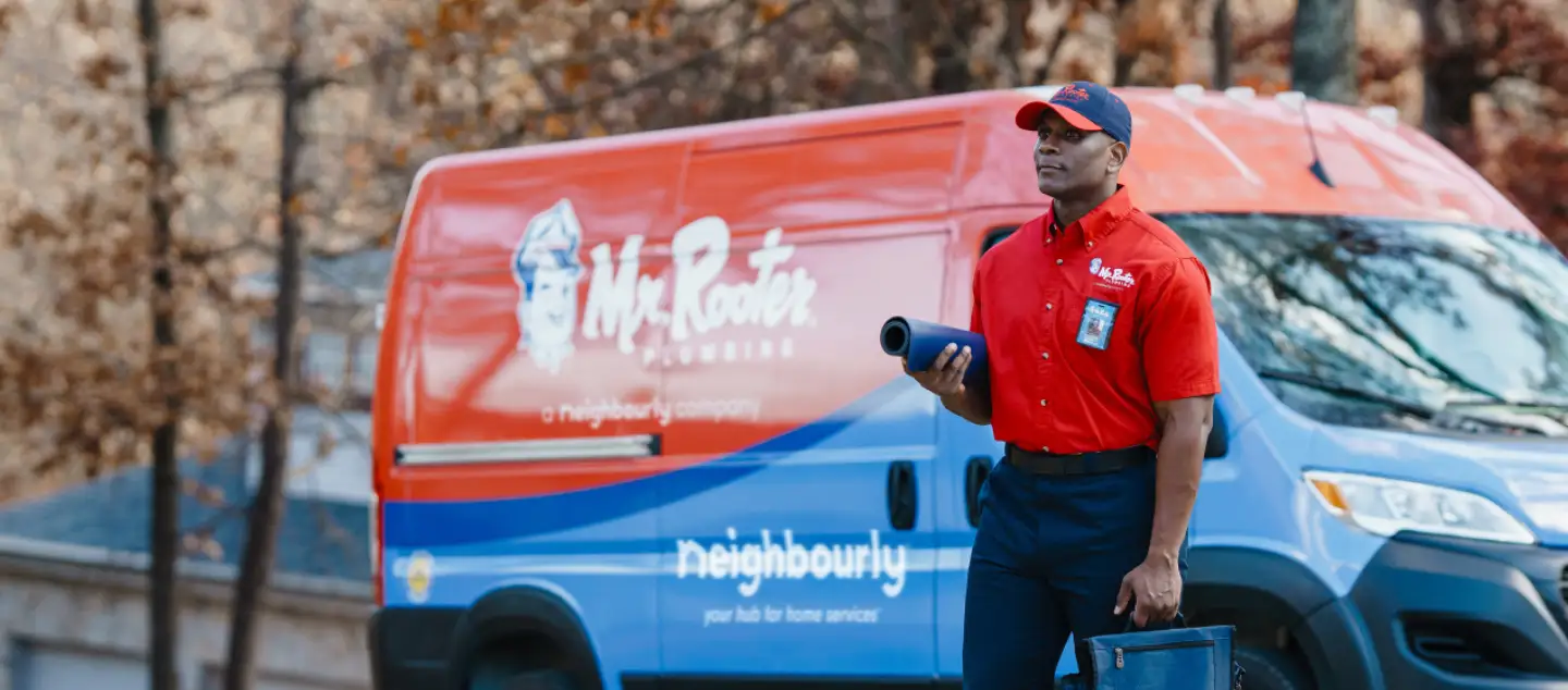 Mr. Rooter technician carrying mat and utility bag in front of branded red and blue work van.