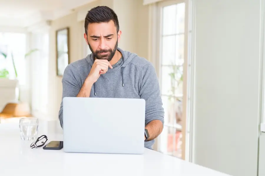  Man in a gray hoodie looks thoughtfully at a laptop while sitting at a table.