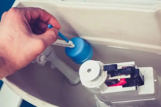 A person pinching the top of a float in an open toilet tank to adjust its height in order to prevent the toilet from running.