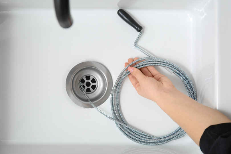Hand using a plumber&rsquo;s drain snake near a clogged bathroom sink during simple DIY plumbing repair in Canada.